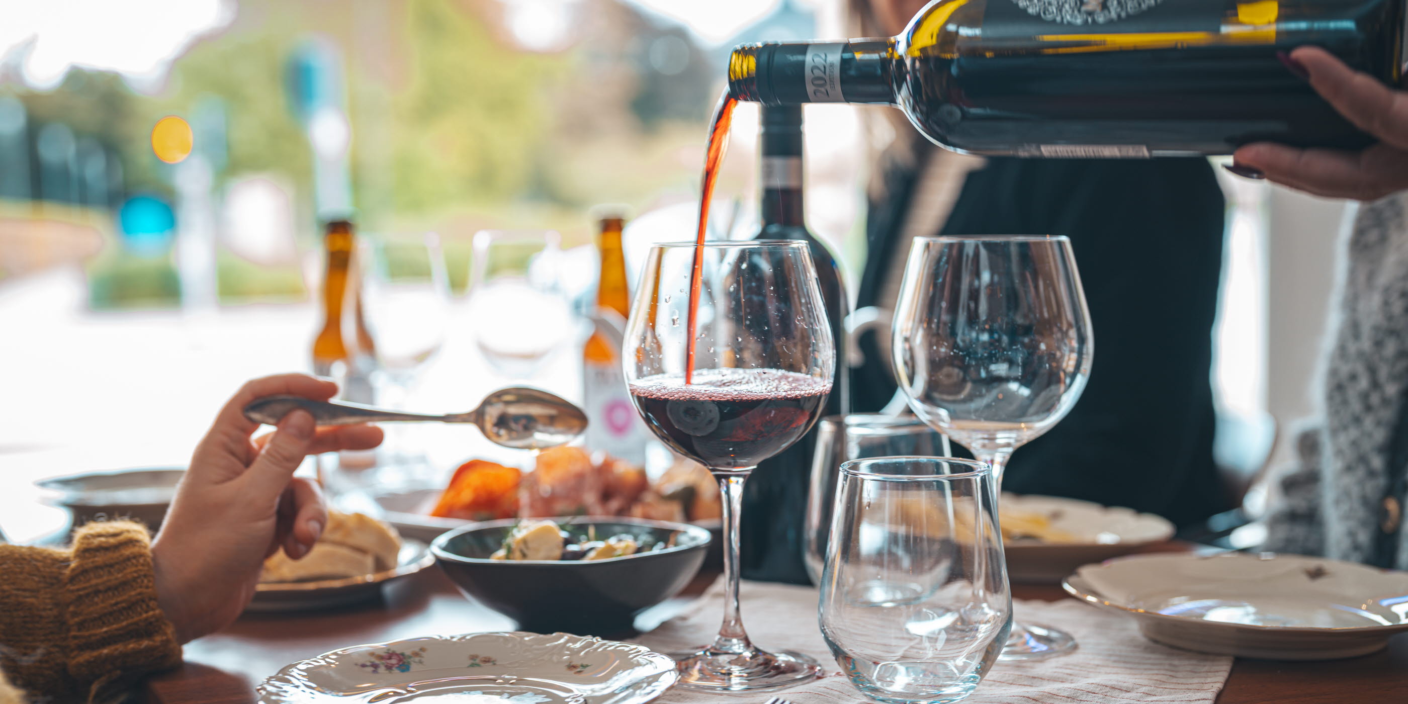 En flaska rödvin hälls upp i ett vinglas vid ett dukat bord med flera tallrikar och glas.